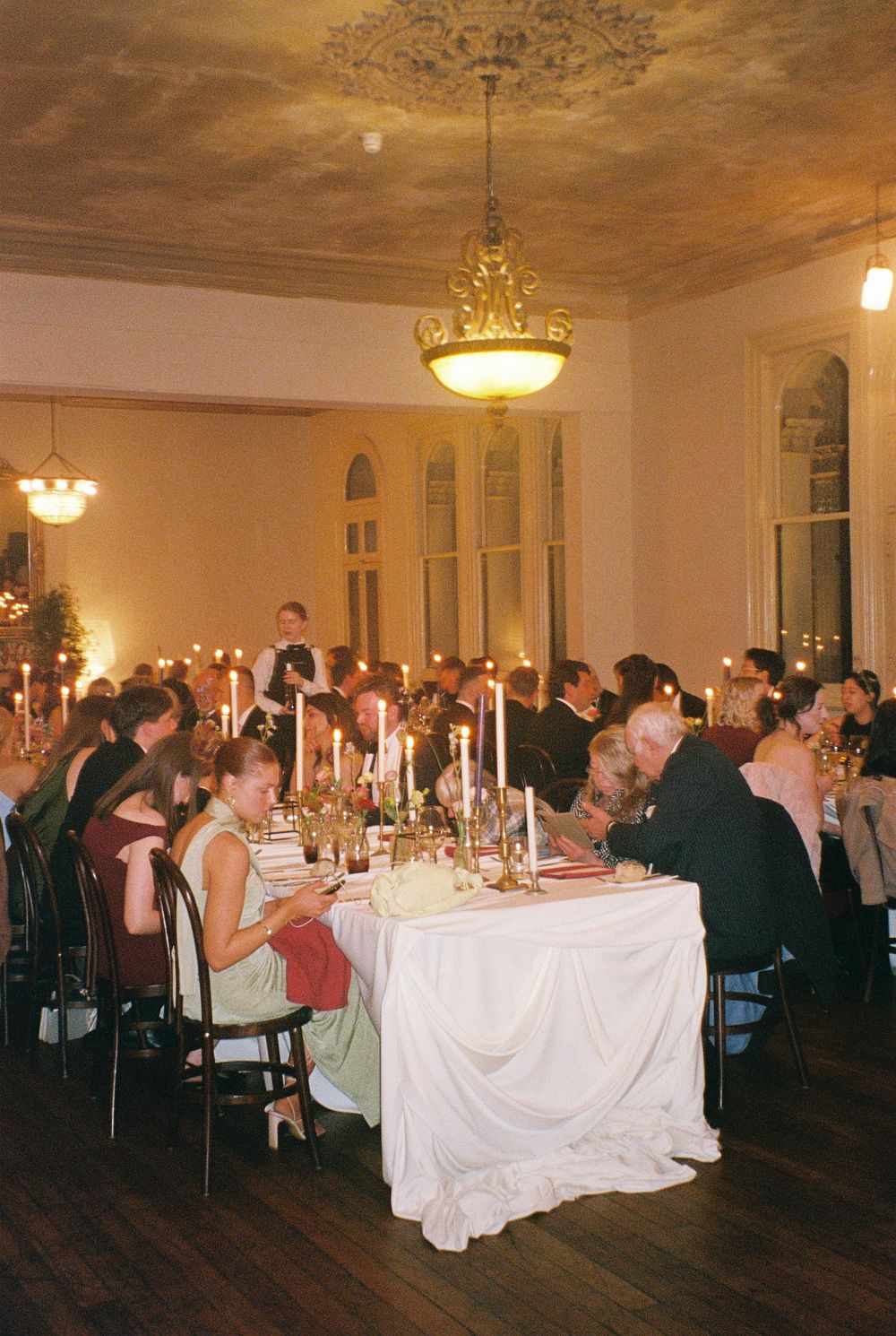 Elegant candlelit wedding reception dinner in a grand ballroom with guests seated at white-draped tables under ornate ceiling.