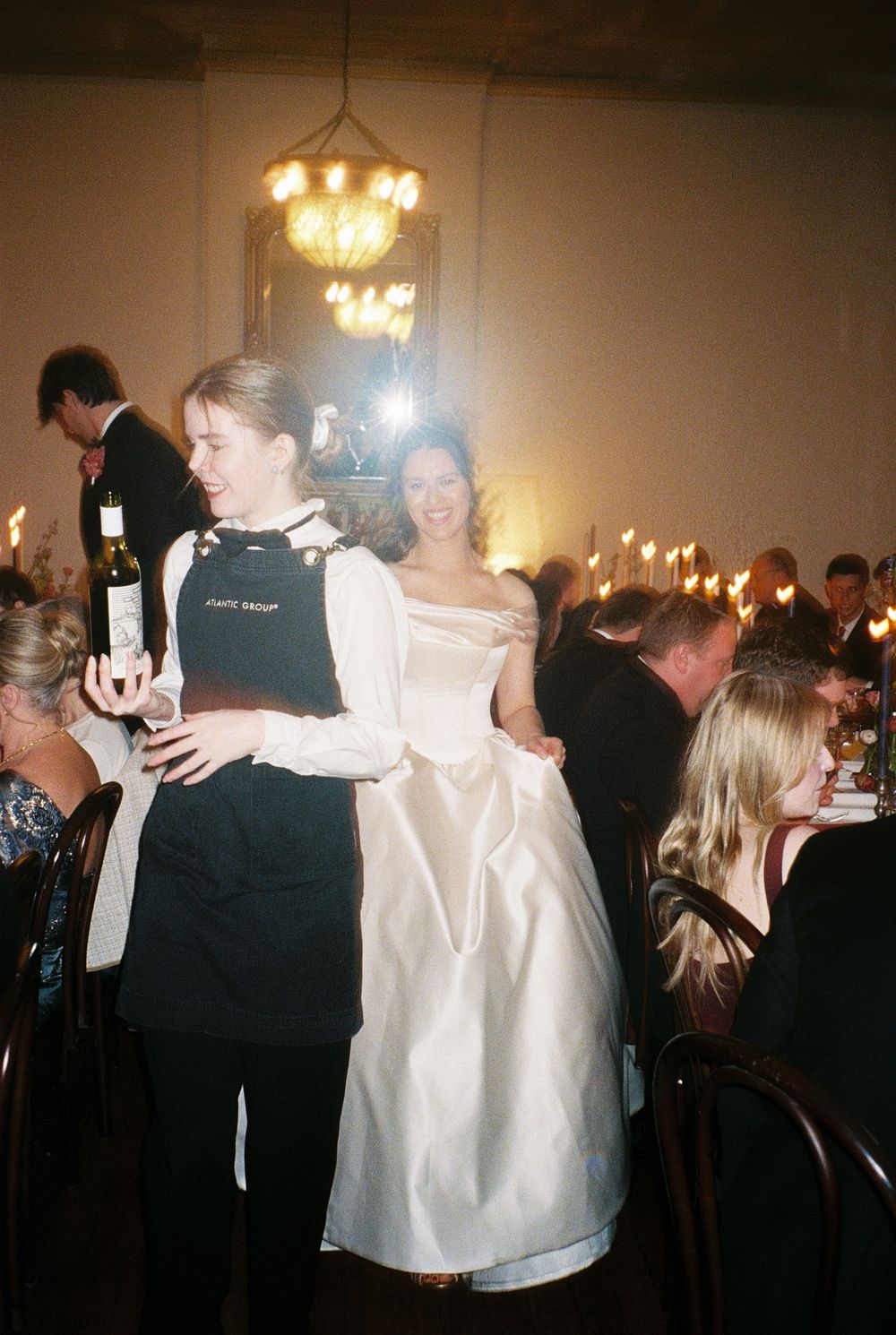 A bride in a white gown stands with a server holding wine at an elegant wedding reception.