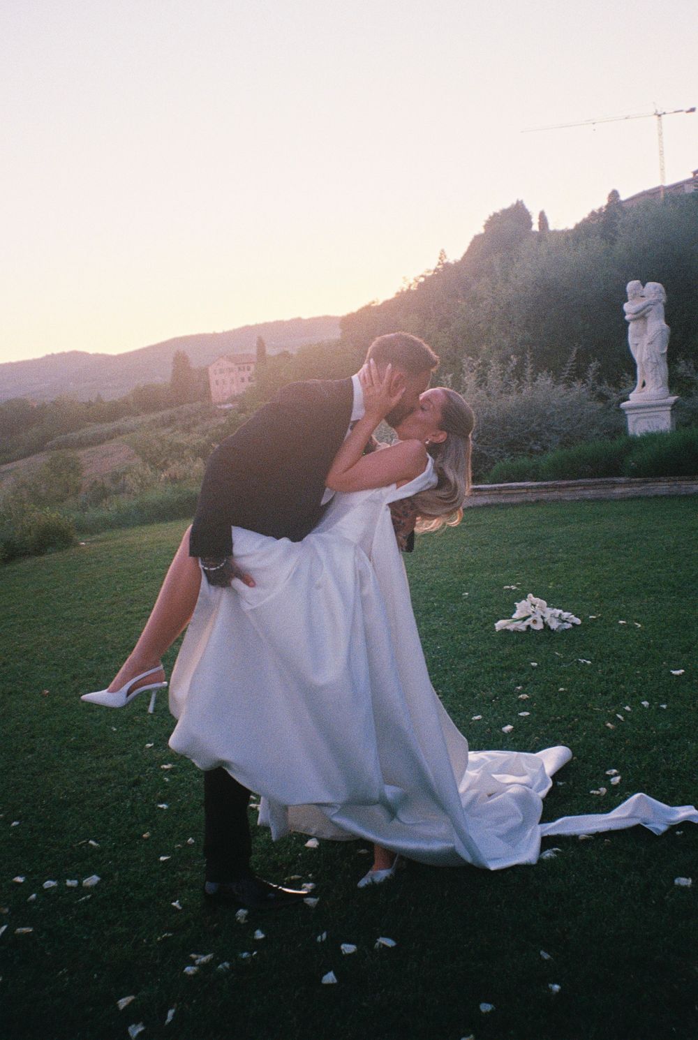 Groom lifts bride in white gown for romantic kiss at sunset, with a garden statue and rolling hills in the background.