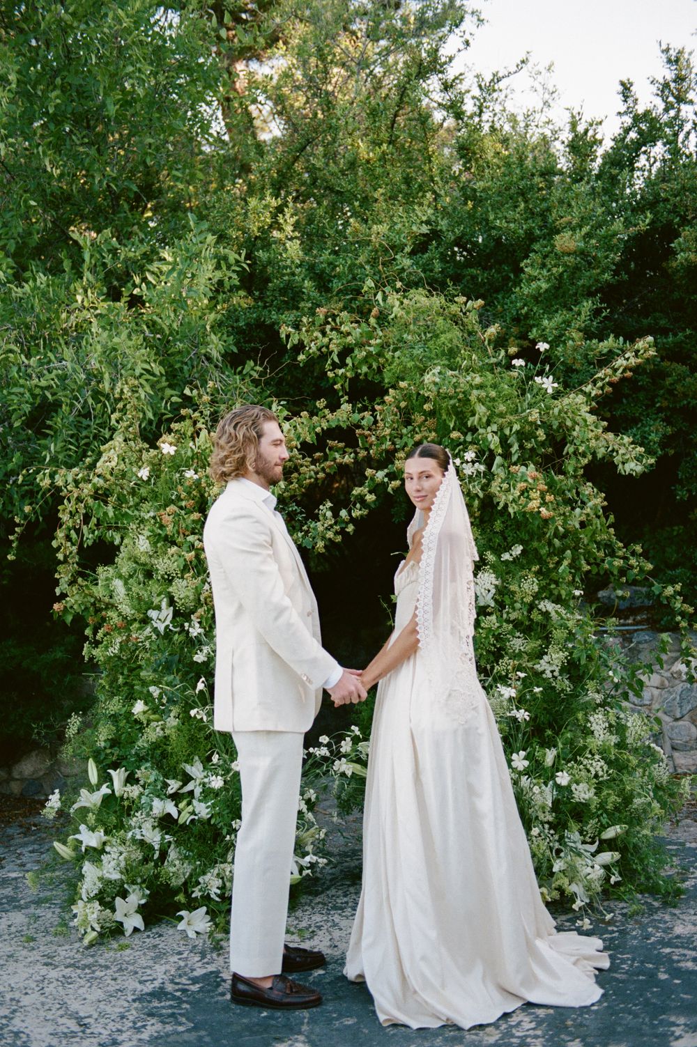 Bride and groom holding hands before a green floral arch with white lilies at outdoor wedding.