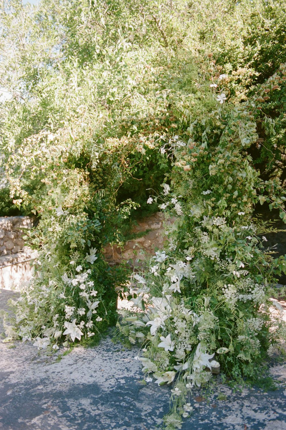 Lush green floral arch adorned with white lilies and cascading foliage for an outdoor wedding ceremony.