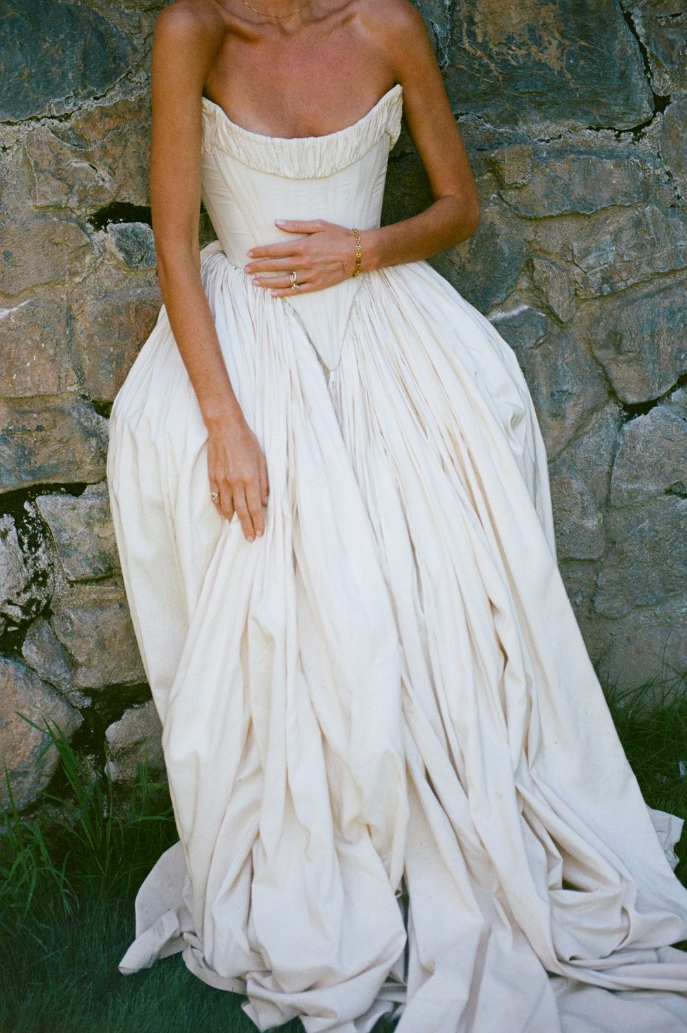 Bride wearing a strapless ivory ball gown wedding dress with ruched bodice, standing against a rustic stone wall.