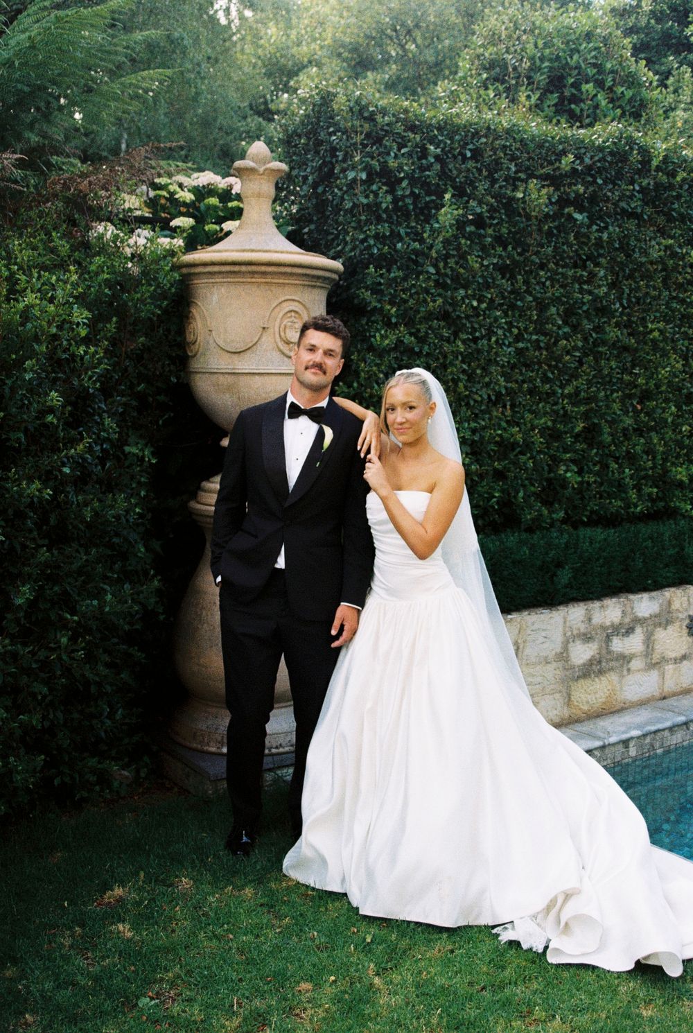 A couple in formal wedding attire pose together in an elegant garden setting with manicured hedges and a pool.