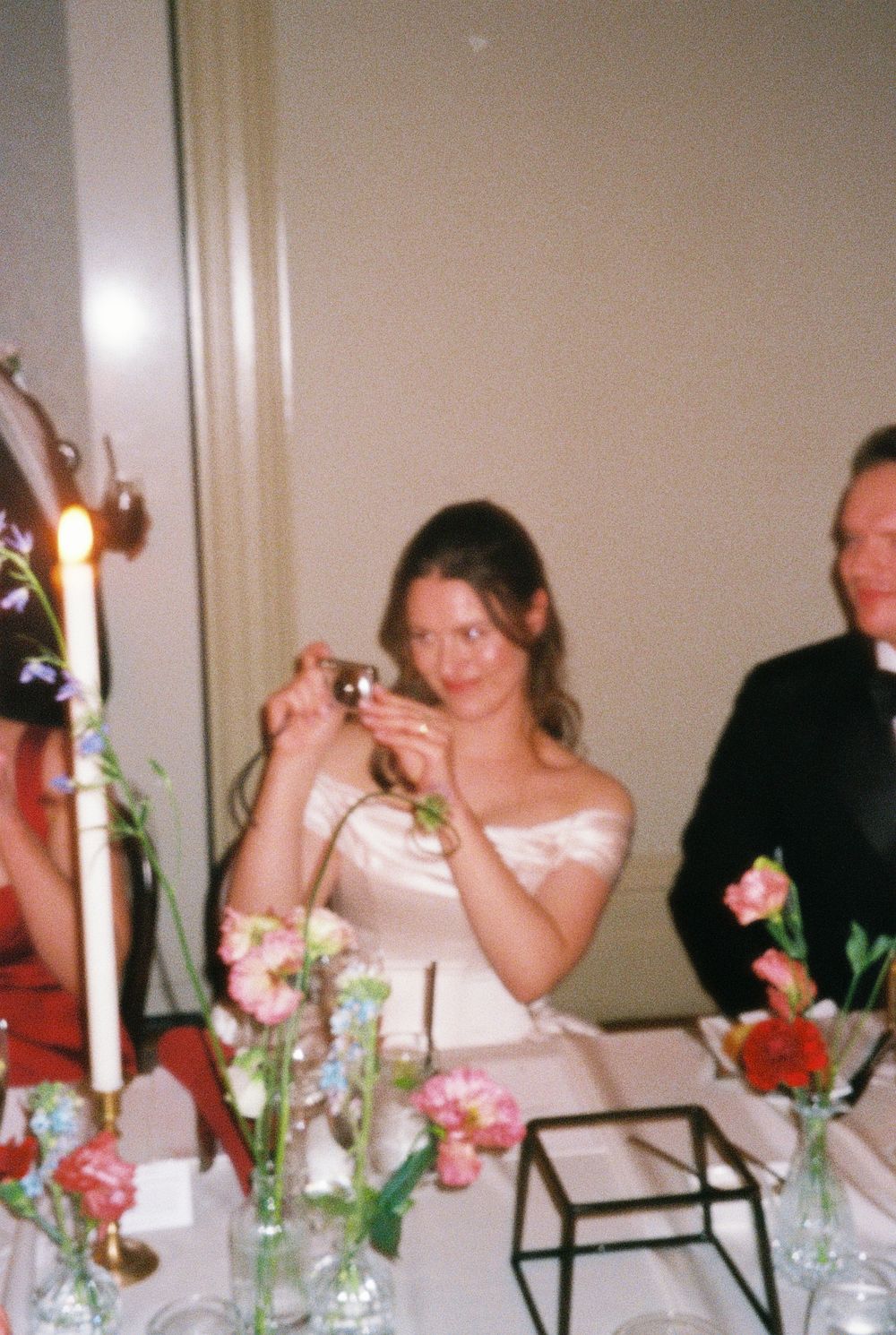 Bride in white strapless gown photographs guests at elegant wedding reception with candles and floral centerpieces.