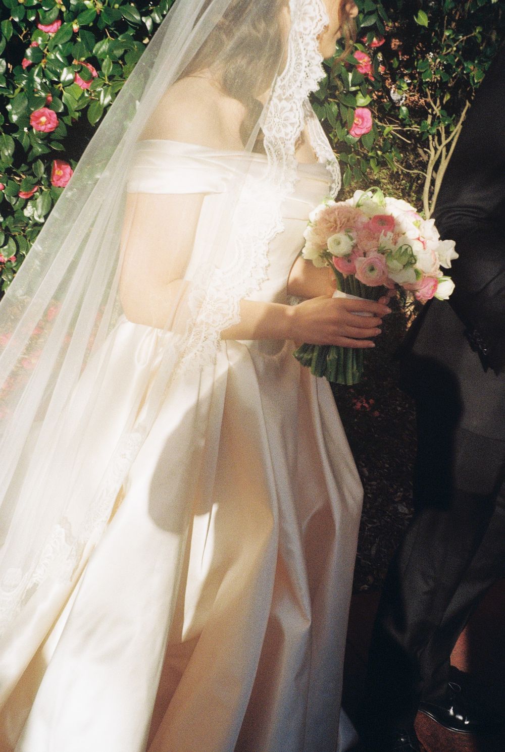 Bride in elegant off-shoulder white wedding gown with lace veil holding a pink and white floral bouquet outdoors.