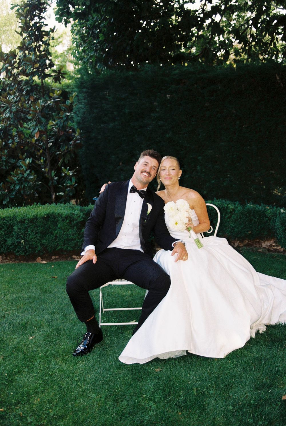 A couple in wedding attire lounges playfully on the grass in a garden setting.