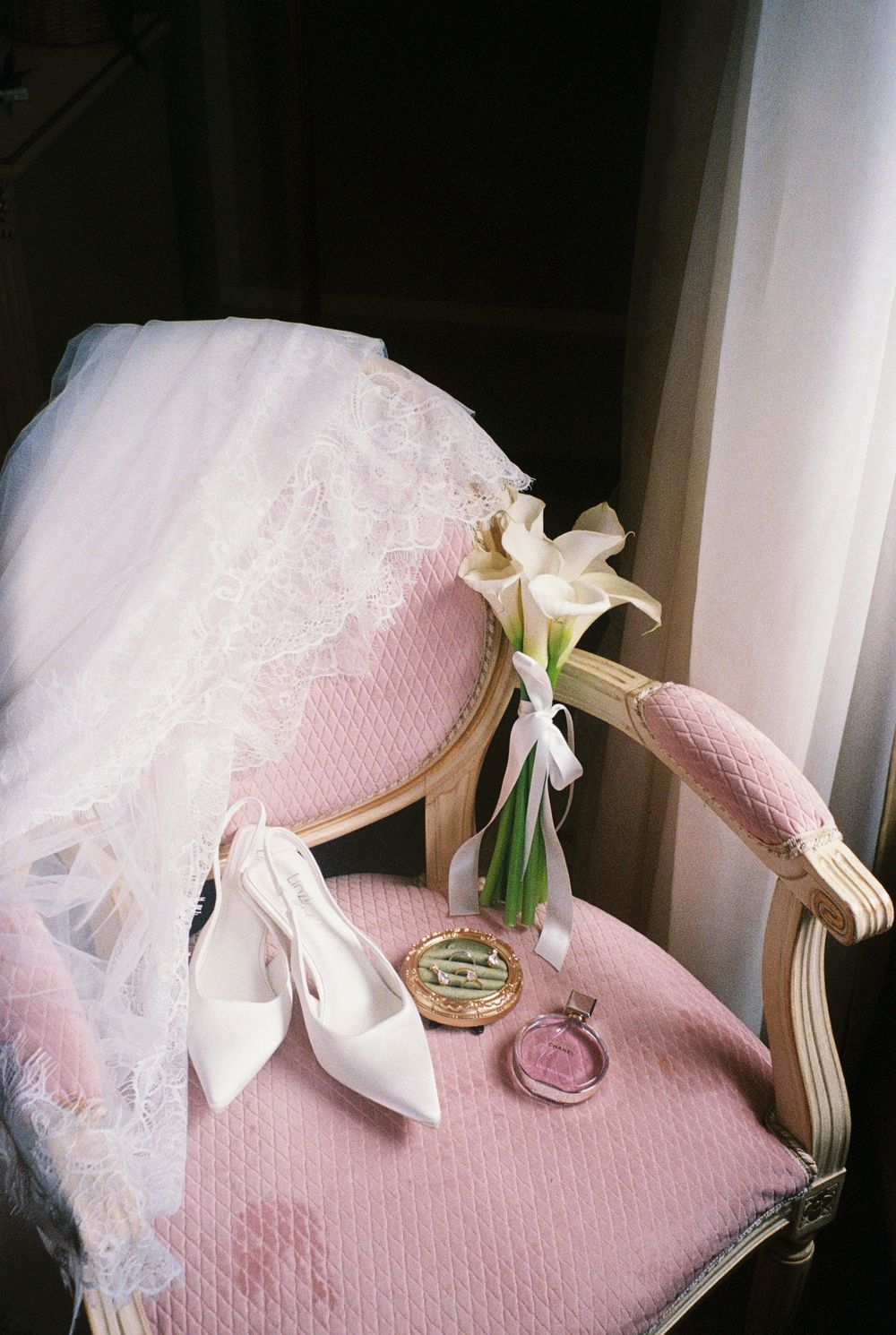 Elegant bridal details on pink chair: white heels, calla lily bouquet, lace veil, and vintage compact mirror.