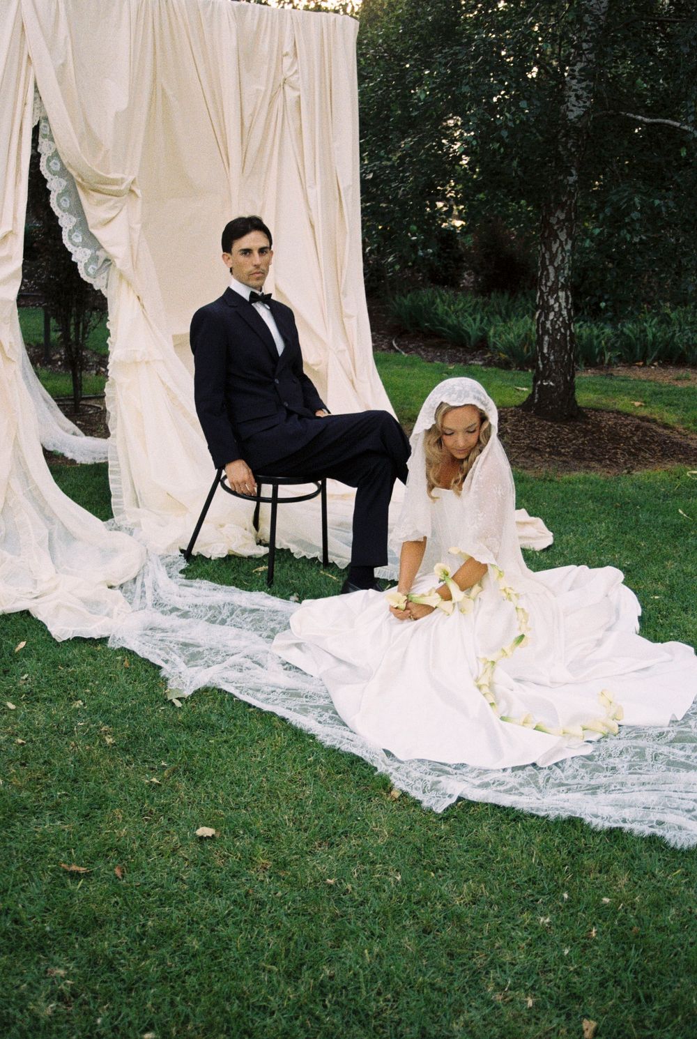 A couple poses for a wedding photo on white fabric draped outdoors, with the groom seated on a chair and bride on the ground.