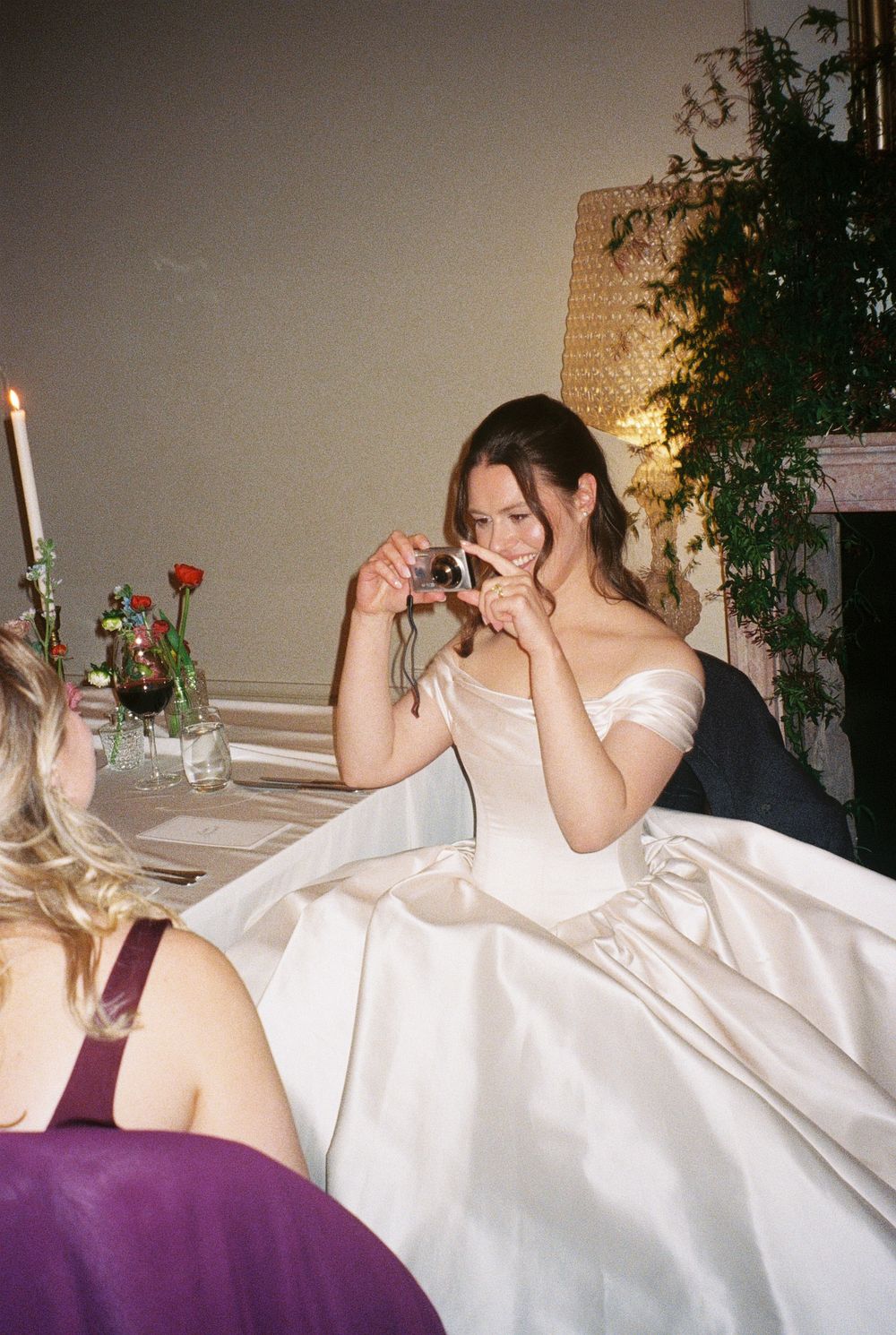 Bride in white off-shoulder ball gown taking photo with camera at elegant wedding reception dinner table.