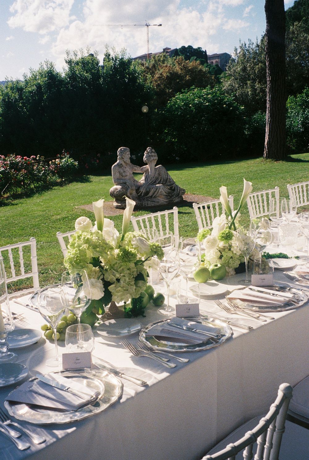 Elegant outdoor wedding reception table with white floral centerpieces, green hydrangeas, and classical stone statues in a lush garden.