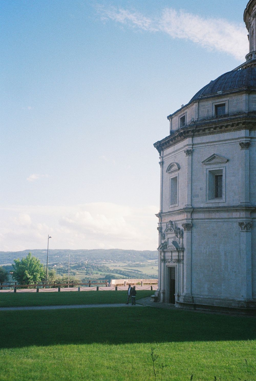 Historic baroque church on a hilltop with lush green lawn and scenic Italian countryside valley view under blue sky.