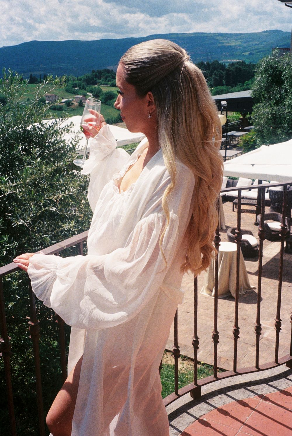 Woman in sheer white robe sipping champagne on a balcony with scenic mountain and vineyard views.