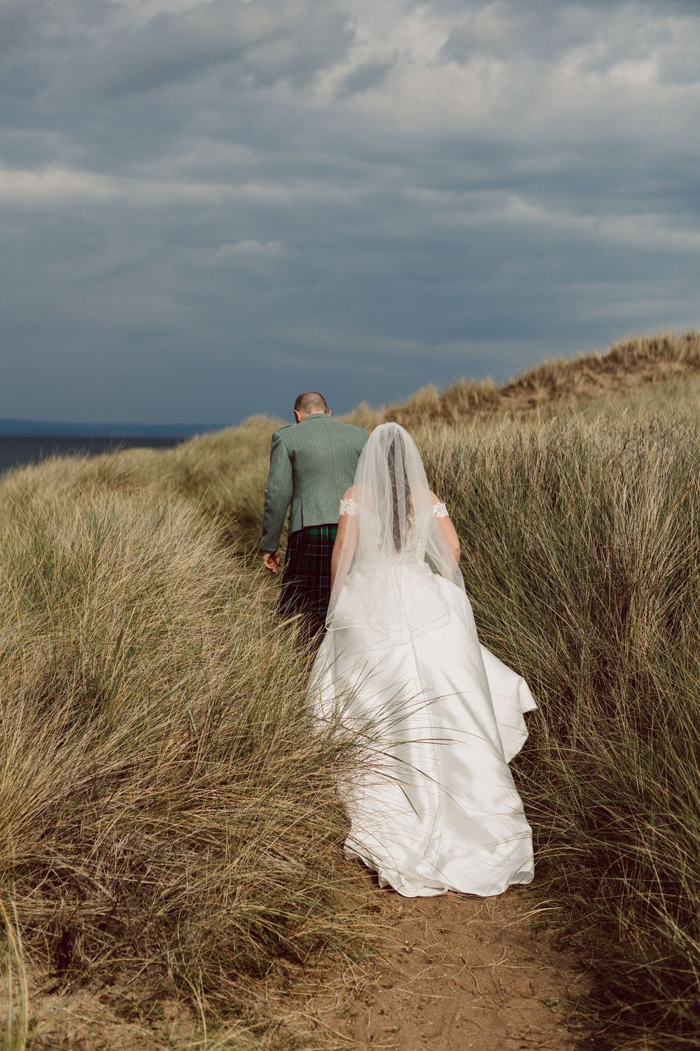 A white wedding dress flows elegantly through coastal sand dunes under moody cloudy skies.