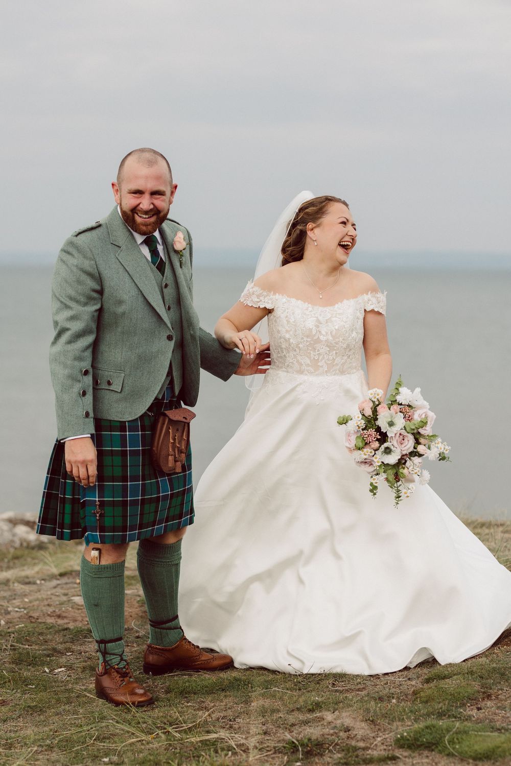 A joyful wedding couple poses together on a coastal cliff wearing traditional Scottish attire and white wedding dress.