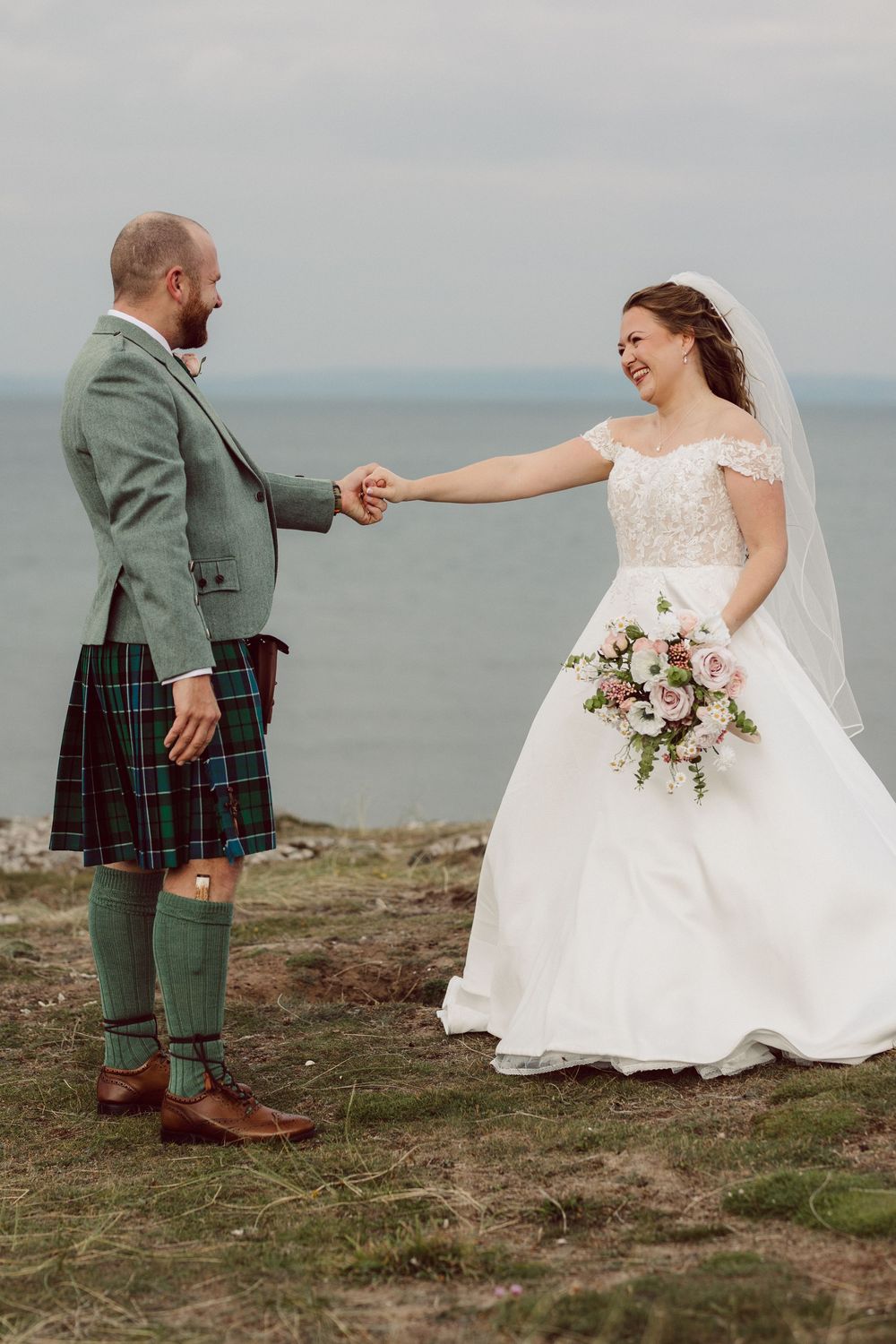A couple in wedding attire poses on a coastal cliff, with the groom wearing a traditional Scottish kilt and green jacket.
