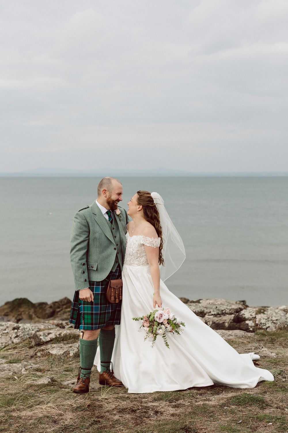 A newlywed couple shares a romantic moment on a seaside cliff, with the groom in traditional Scottish dress and bride in white.