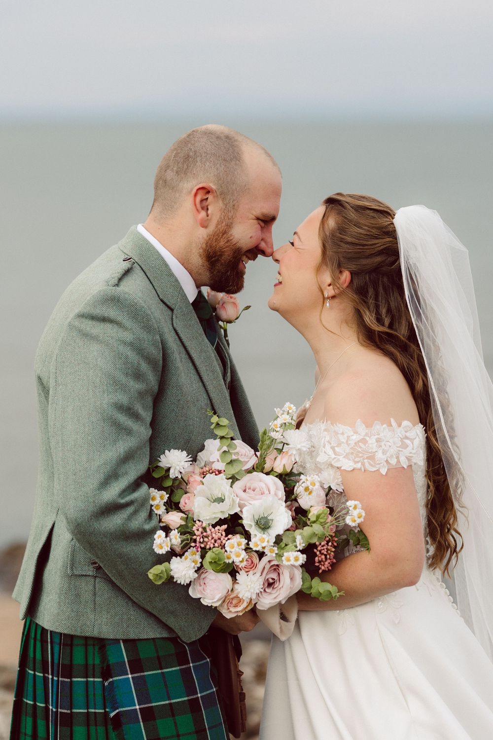 A romantic wedding couple in Scottish attire share tender moments by the sea, with the groom in a green jacket and kilt.