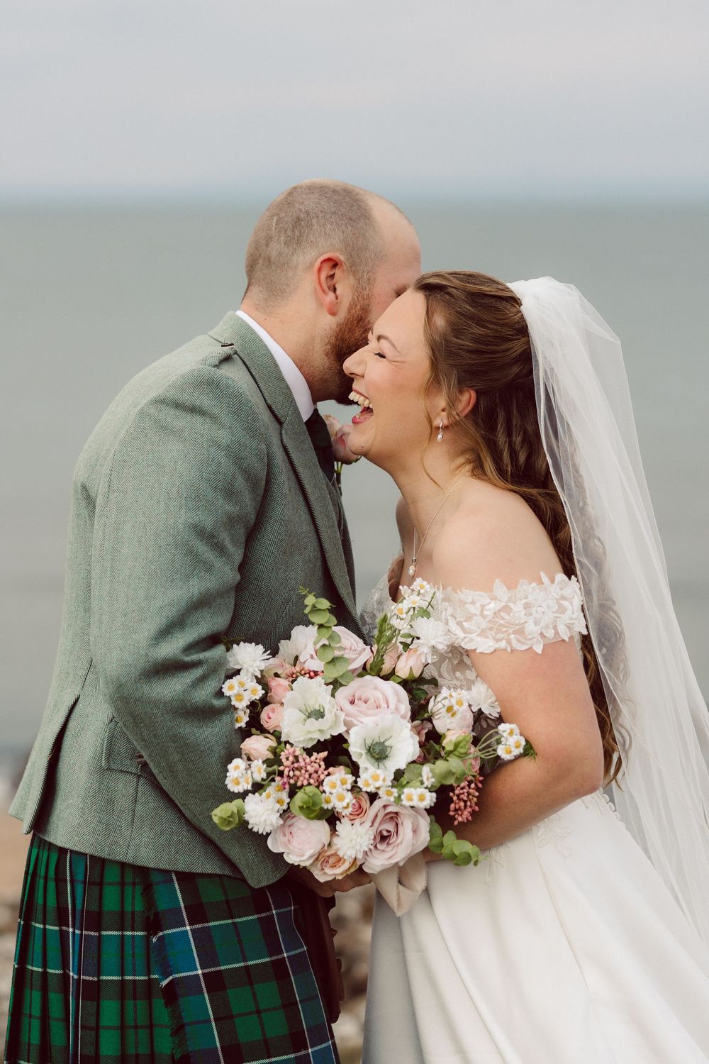 A romantic coastal wedding scene features Scottish tartan and delicate pink floral bouquet against ocean backdrop.