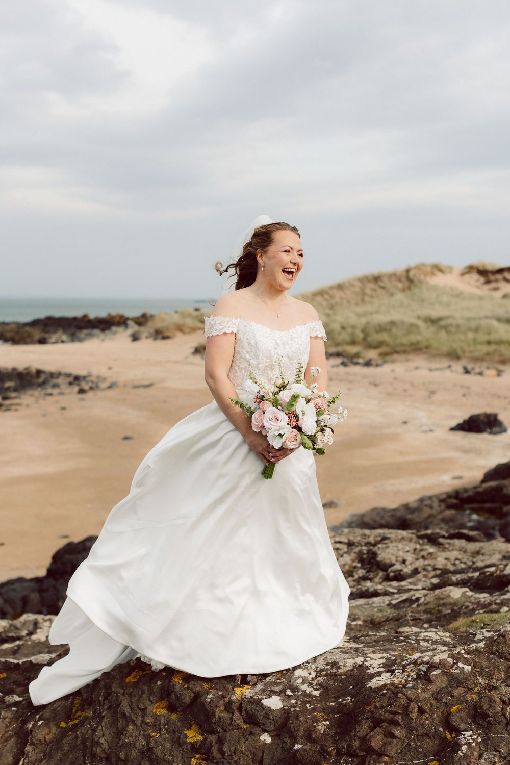 A bride in a flowing white dress sits joyfully on coastal rocks with a bouquet against a cloudy beach landscape.