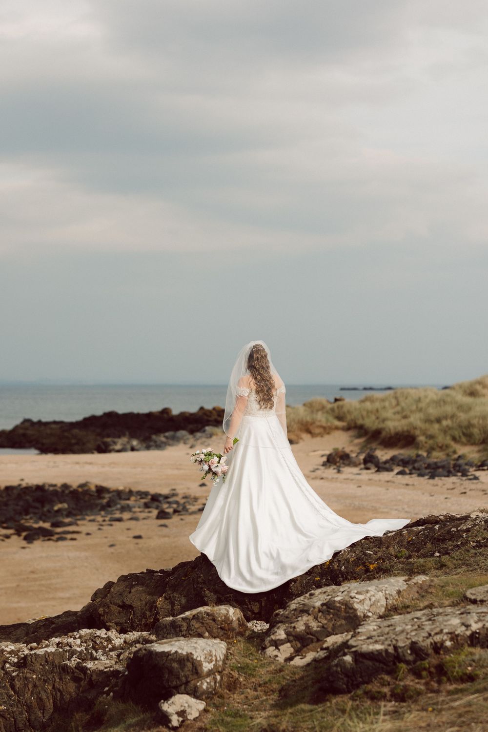 A bride in flowing white gown sits gracefully on rocky coastal cliffs overlooking the sea.
