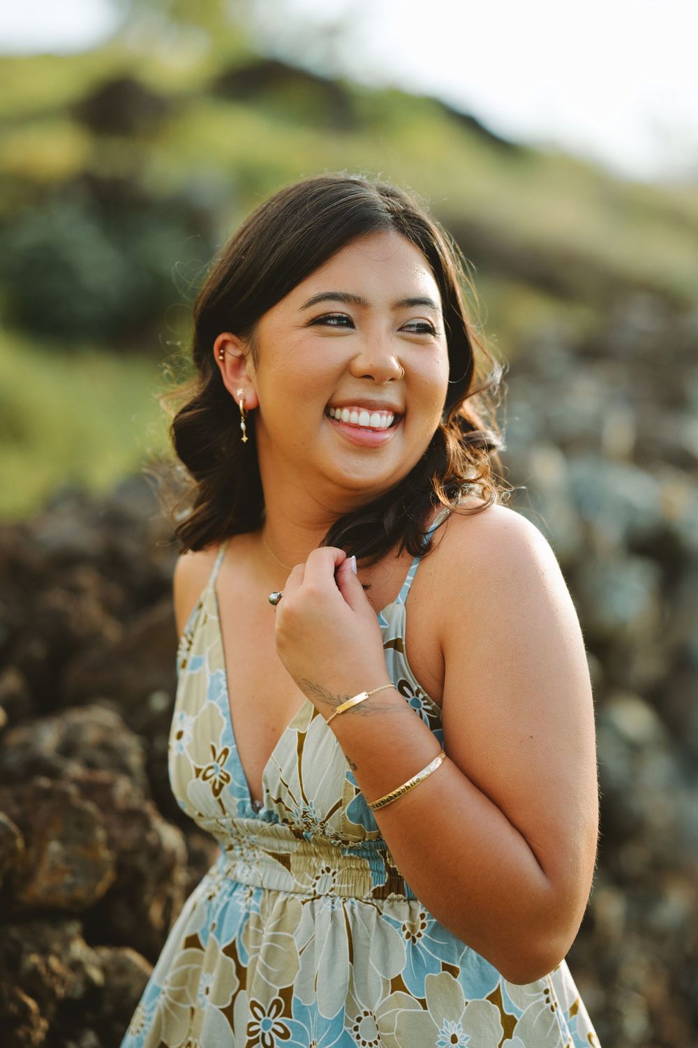 Woman in floral dress smiles brightly against rocky outdoor backdrop during sunset photoshoot.