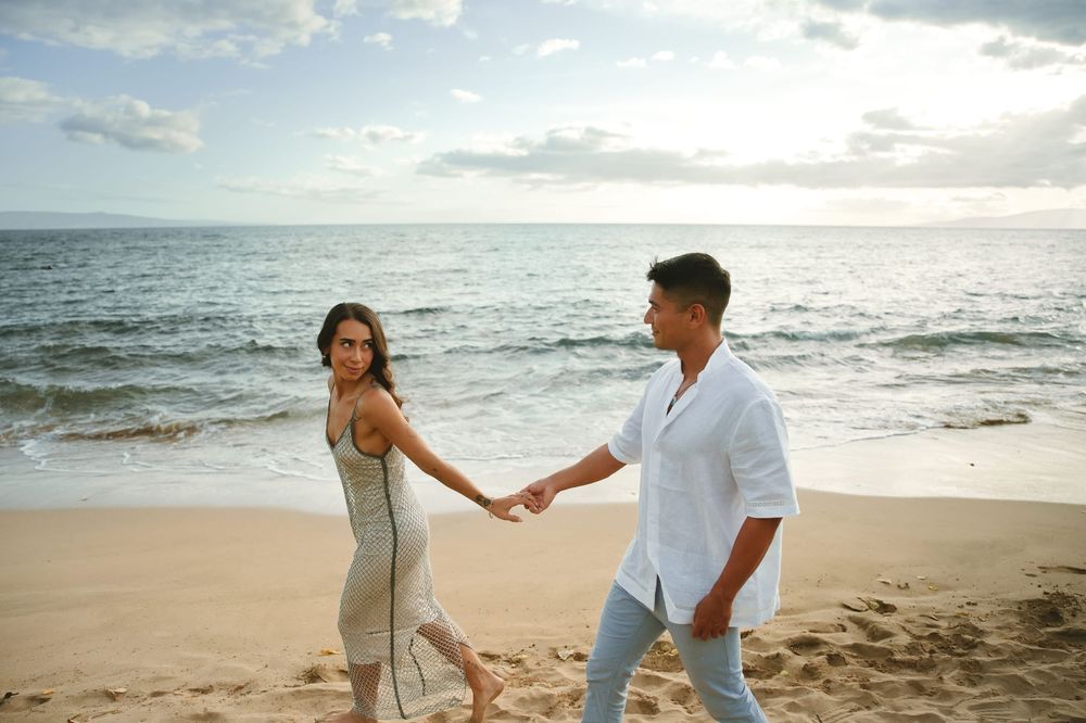 A couple holds hands while walking along a sandy beach at golden hour.