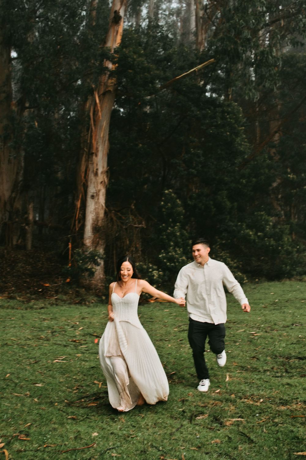 Couple runs playfully through eucalyptus forest wearing coordinated white outfits.