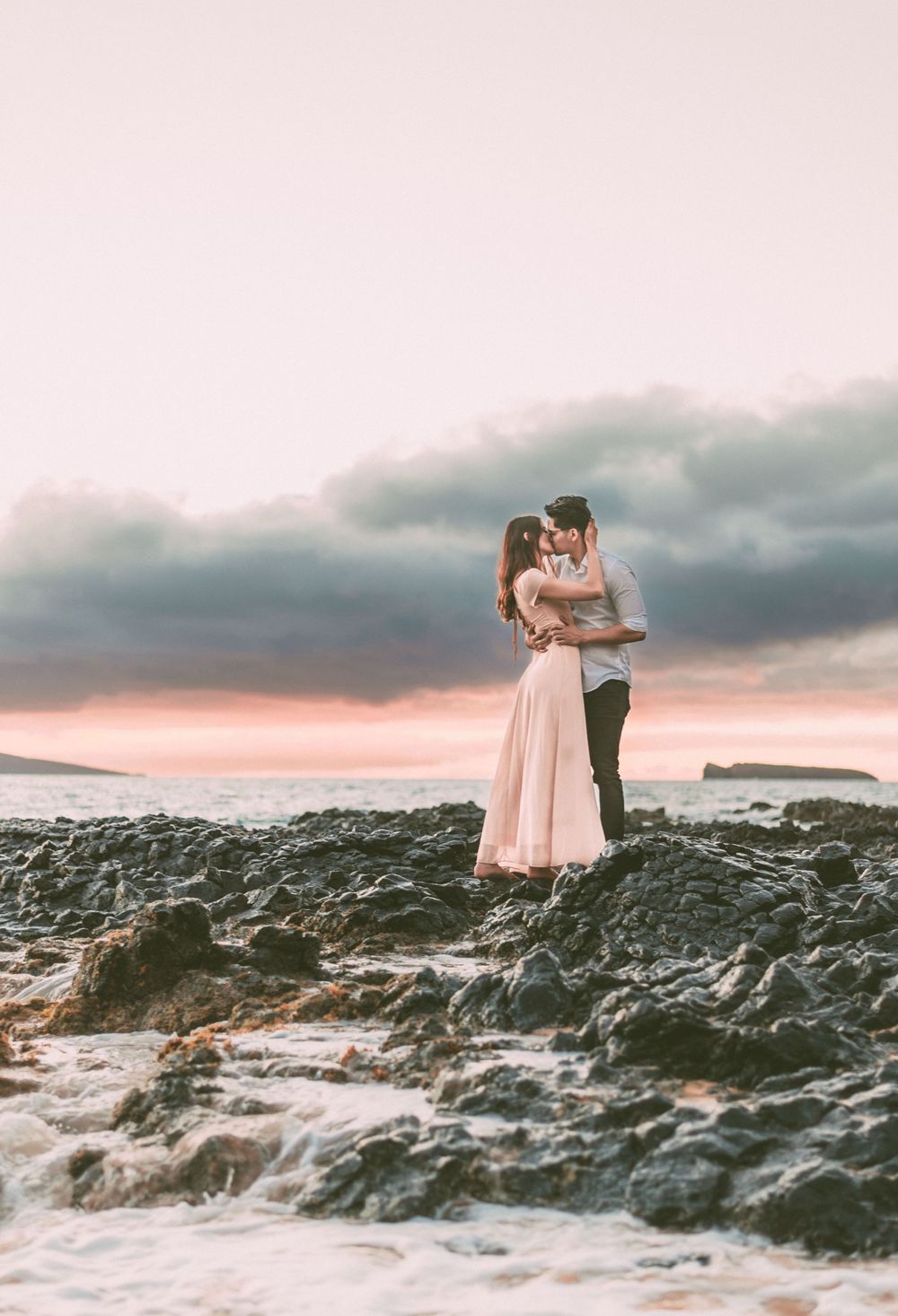 A romantic couple poses on volcanic rocks by the ocean during a pink sunset.