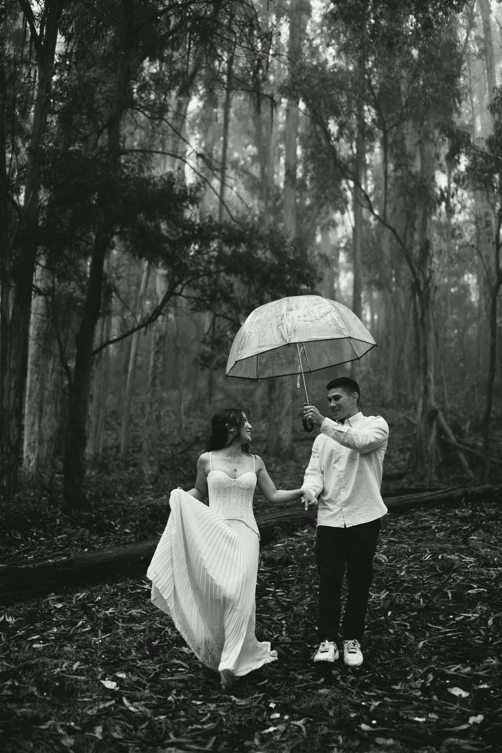 A black and white photo series shows two people in white outfits dancing with umbrellas in a misty forest setting.
