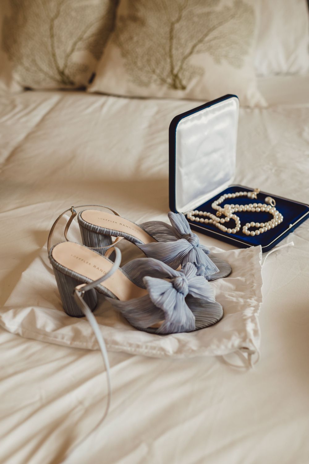 Wedding accessories including gray bow heels and pearl necklace displayed on white bedding.