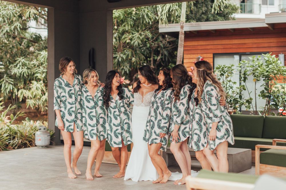 Wedding party gathering under covered outdoor area with tropical greenery in background.