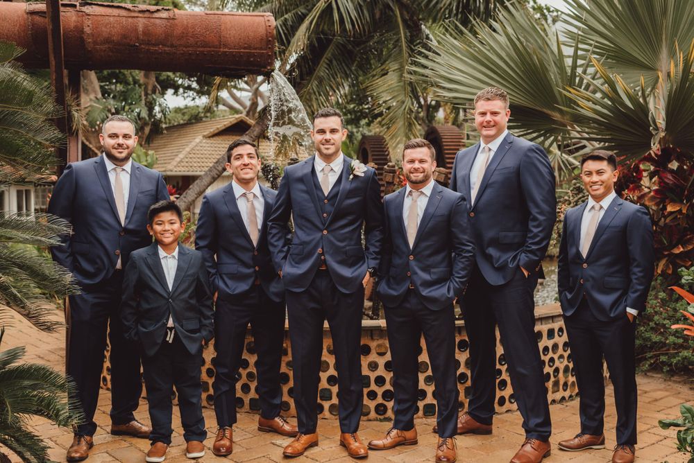Groomsmen in navy suits lined up for formal wedding photos at tropical garden venue.