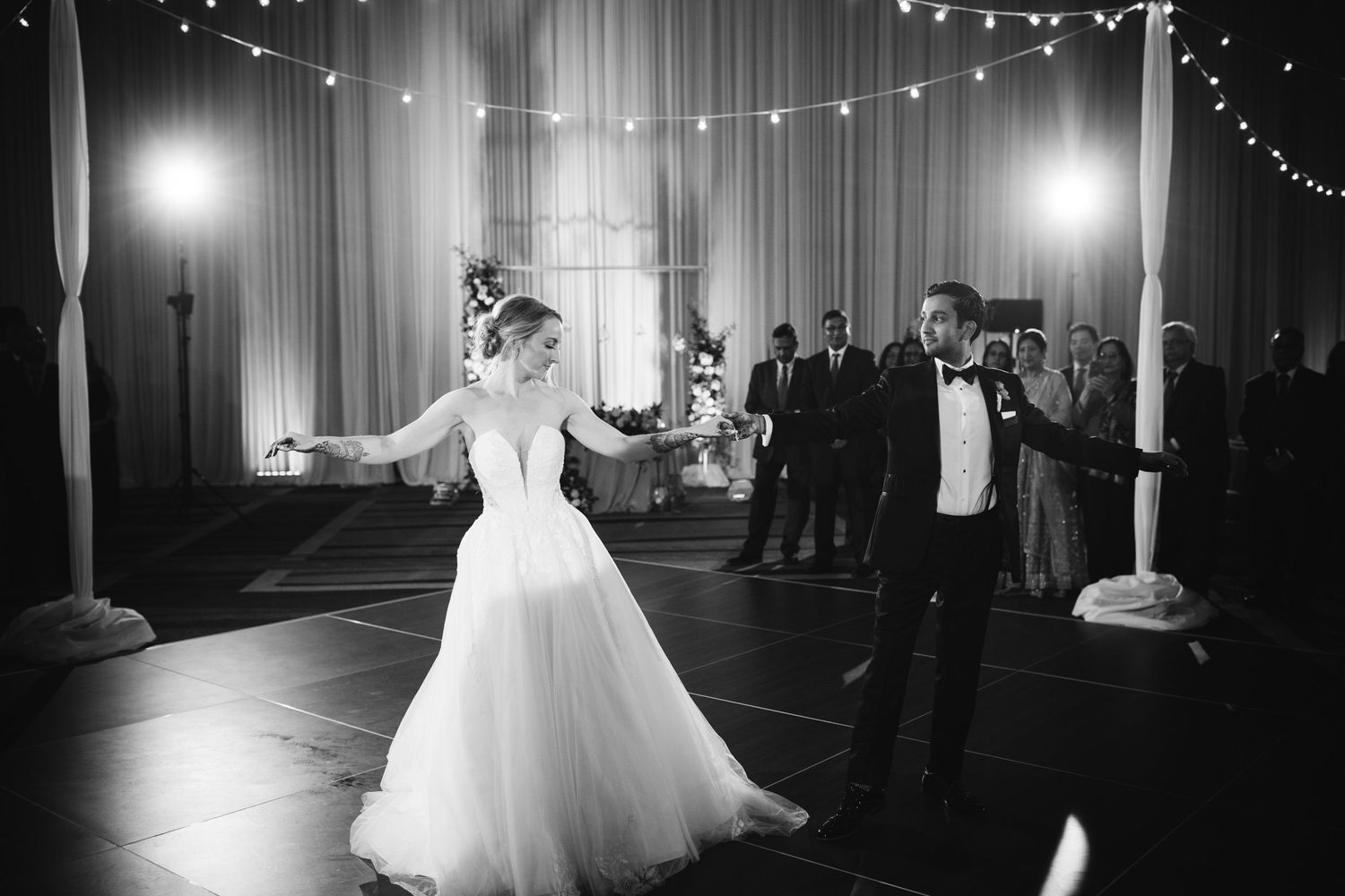A black and white wedding dance sequence showing a couple twirling across a dimly lit reception hall with string lights.