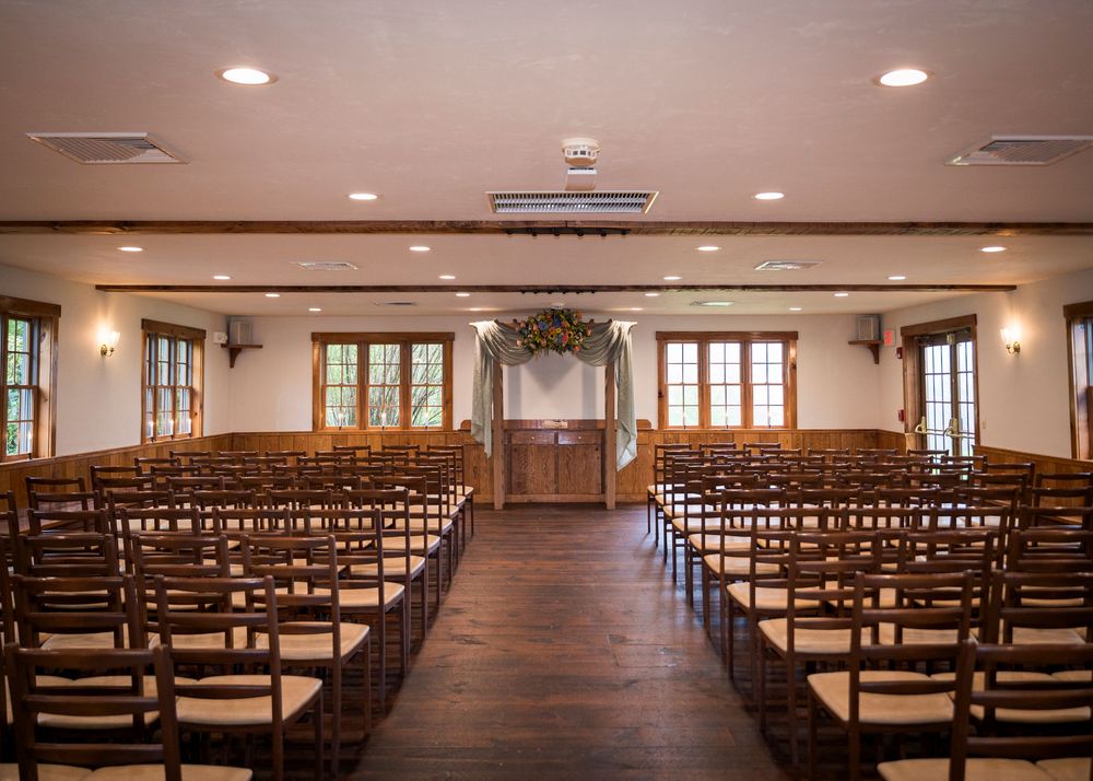 Elegant wedding ceremony venue with wooden chairs, floral arch, and rustic decor in a warmly lit indoor hall.