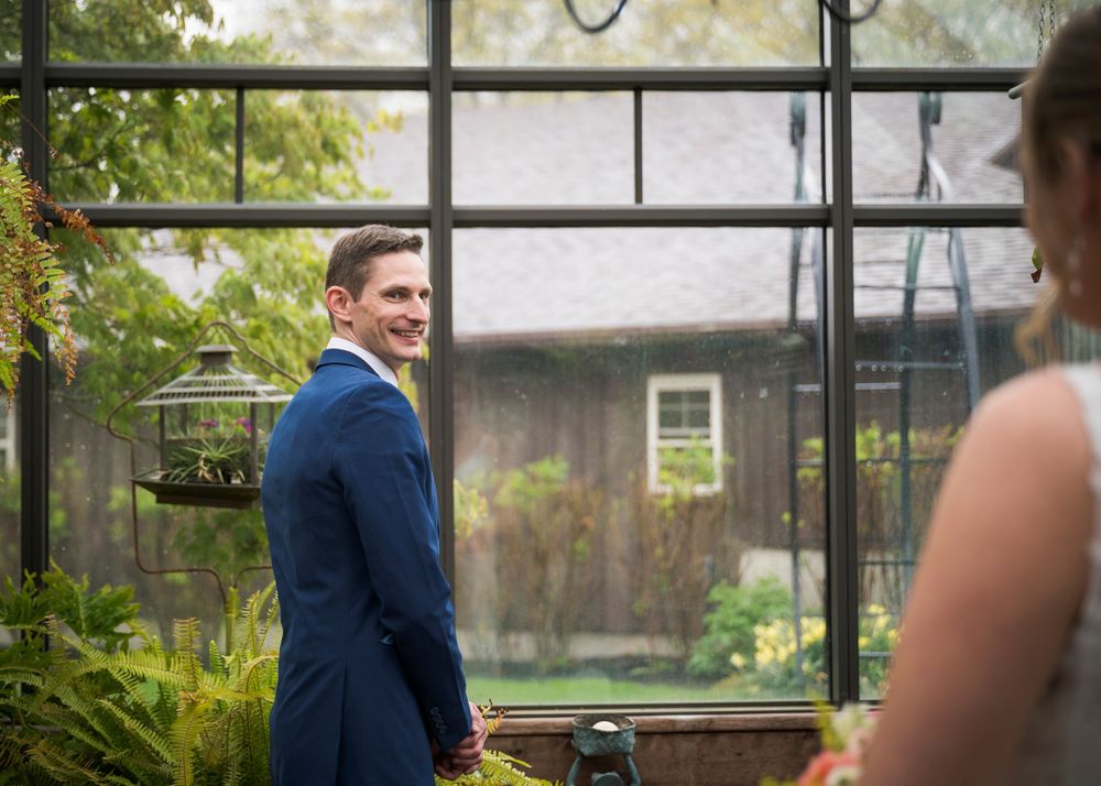 Groom in navy blue suit smiling during first look moment in greenhouse setting.