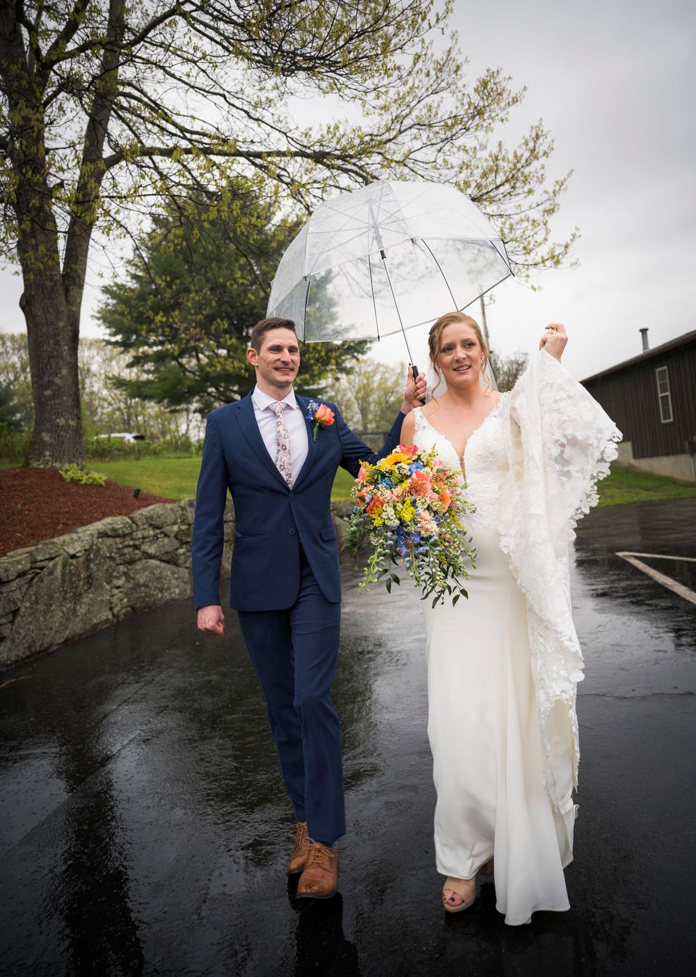 Bride and groom walk together under a clear umbrella on a rainy wedding day, bride holding colorful bouquet.