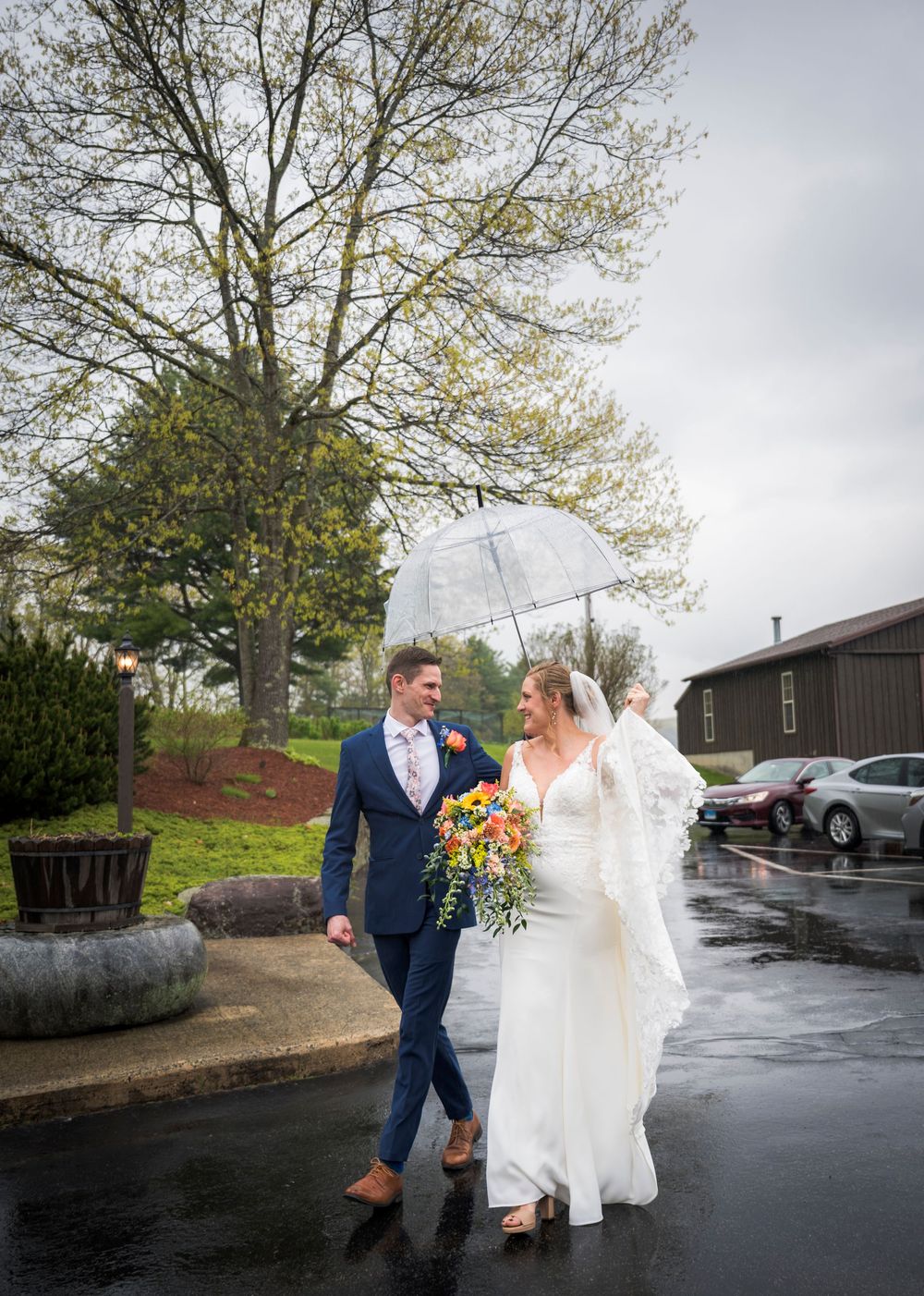 Bride and groom walking under a clear umbrella on a rainy wedding day, bride holding colorful bouquet.