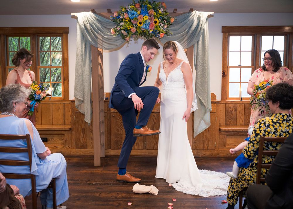 Groom stomping glass during Jewish wedding ceremony while bride watches in elegant white gown.