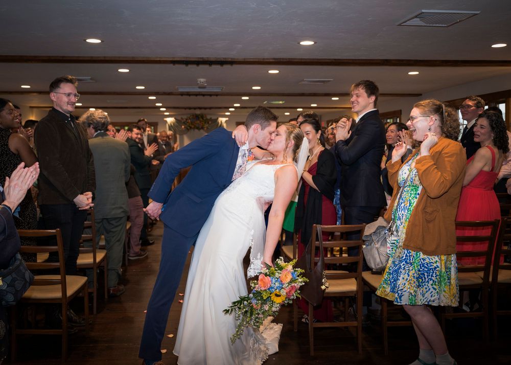 Bride and groom share a kiss while walking down the aisle as guests cheer and applaud at their wedding.