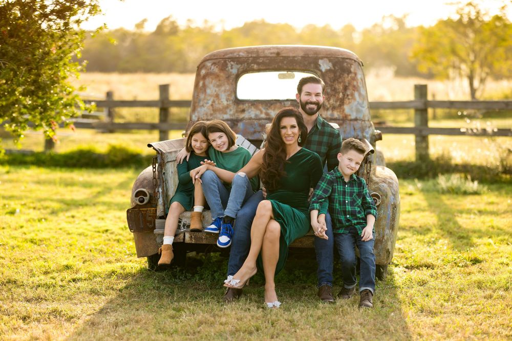 A group sits together on a rustic vintage truck tailgate in a sunlit field at golden hour with wooden fencing in background.