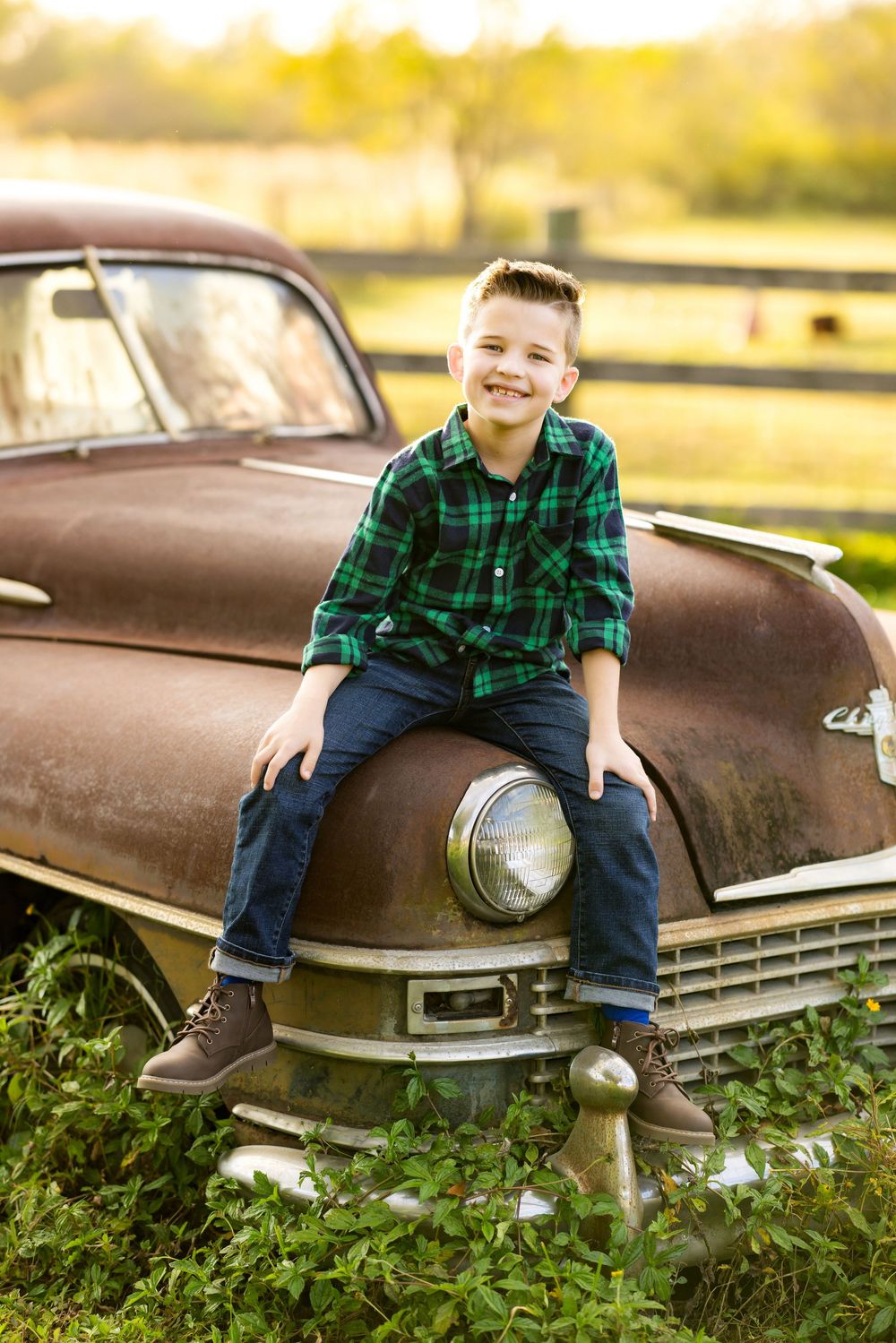A rustic vintage brown car sits in a golden-lit field with a green plaid shirt and jeans visible on the hood.