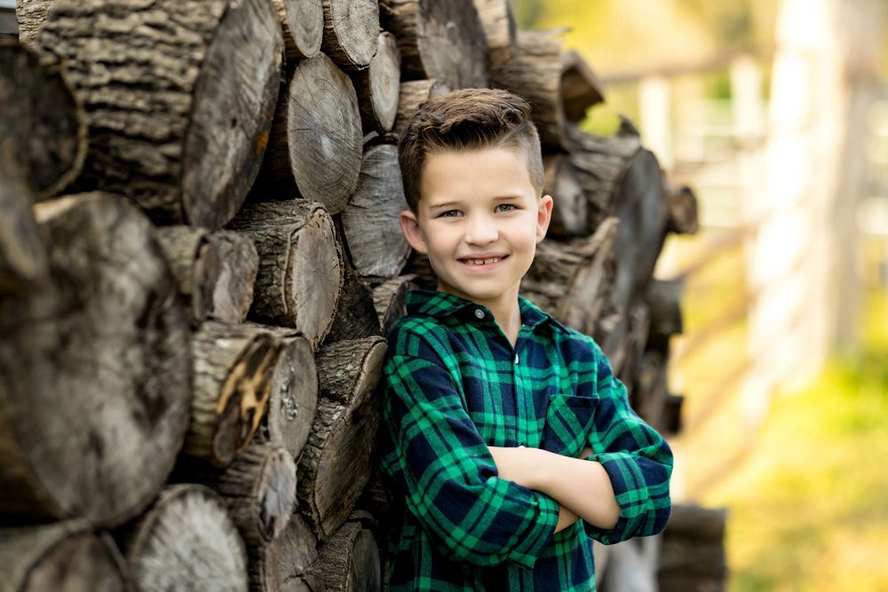 A child in a green plaid shirt poses against a blurred outdoor background in warm sunlight.