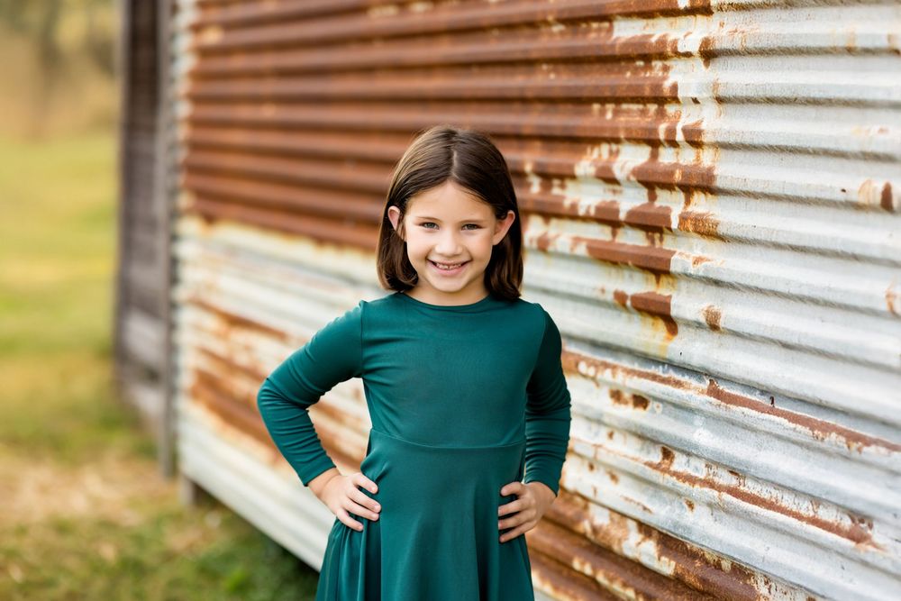 A person in a teal sweater poses against a rustic corrugated metal wall in warm outdoor lighting.