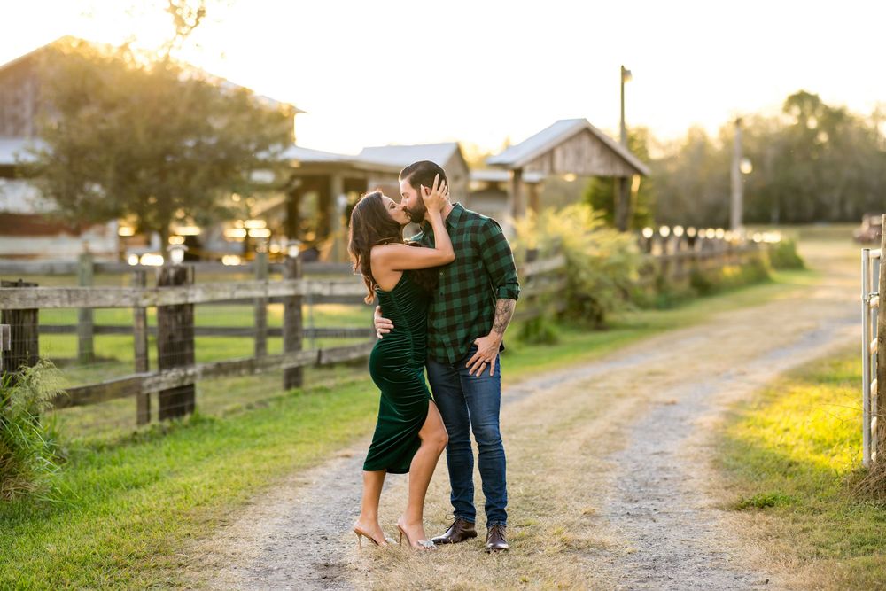 A couple embraces on a dirt path at sunset with a rustic farm and wooden fence in the background.