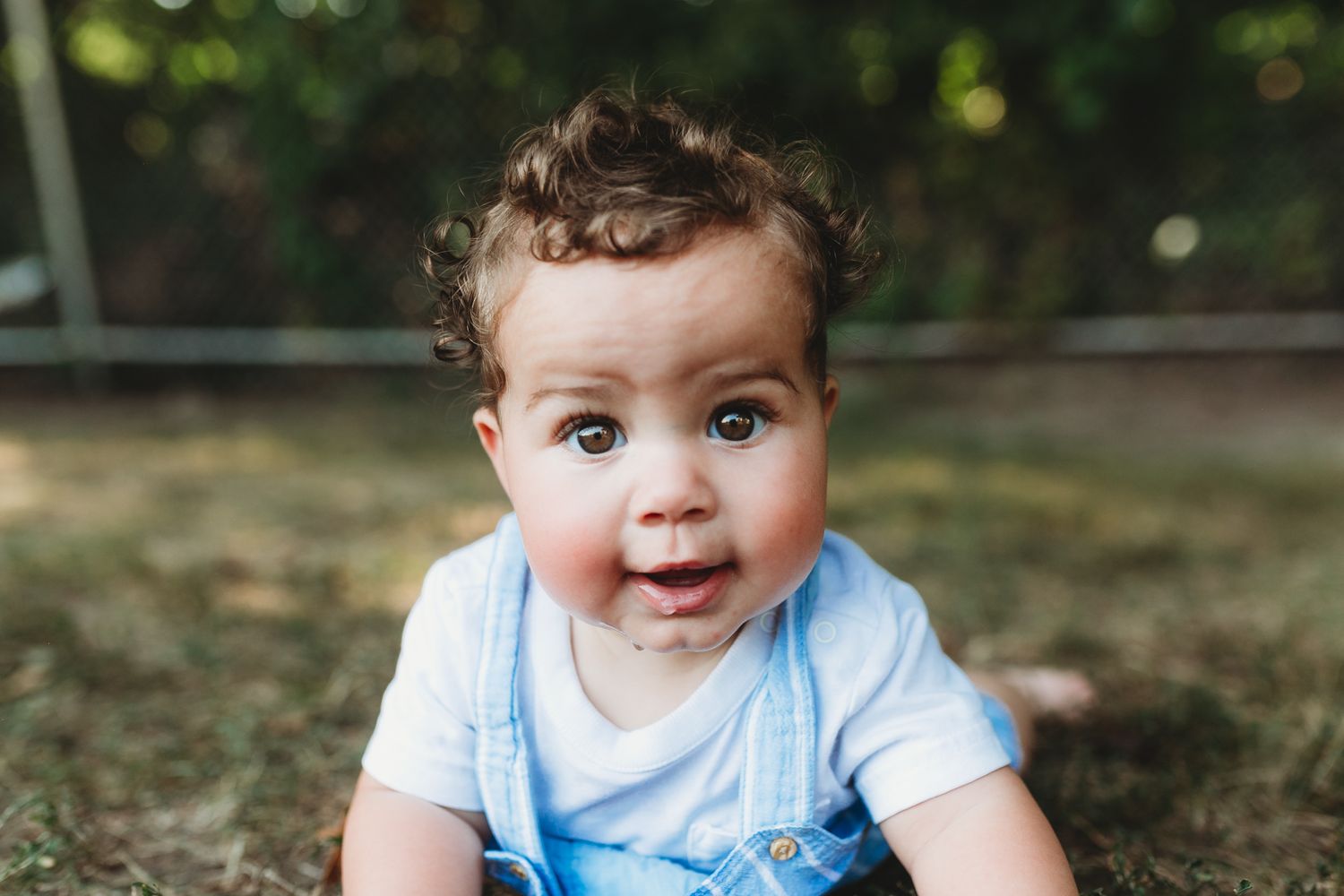 A smiling baby in a light blue outfit sits outdoors against a blurred natural background. Branding kid's clothing. commercial photography