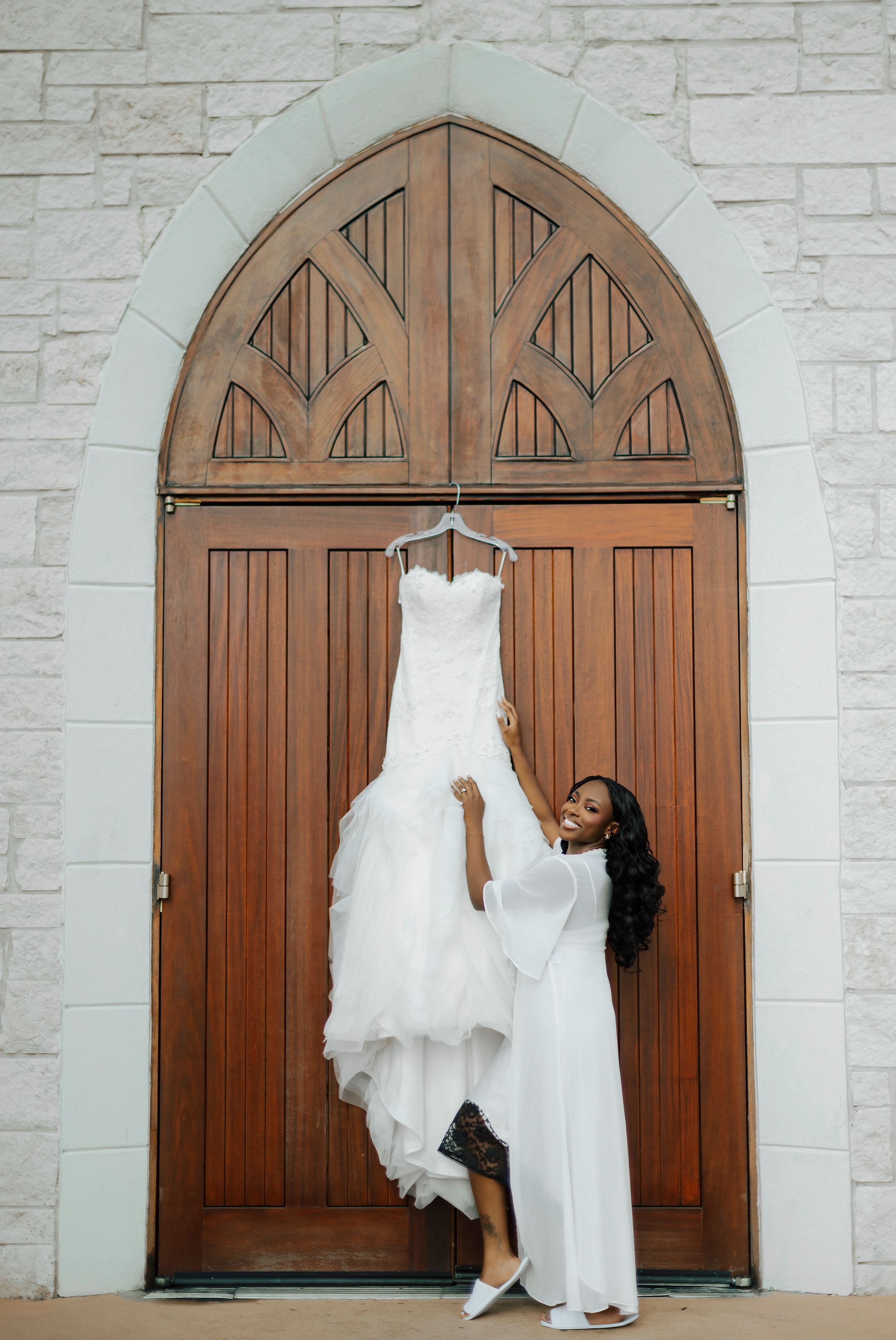 A white wedding gown is displayed with movement against rustic wooden double doors.