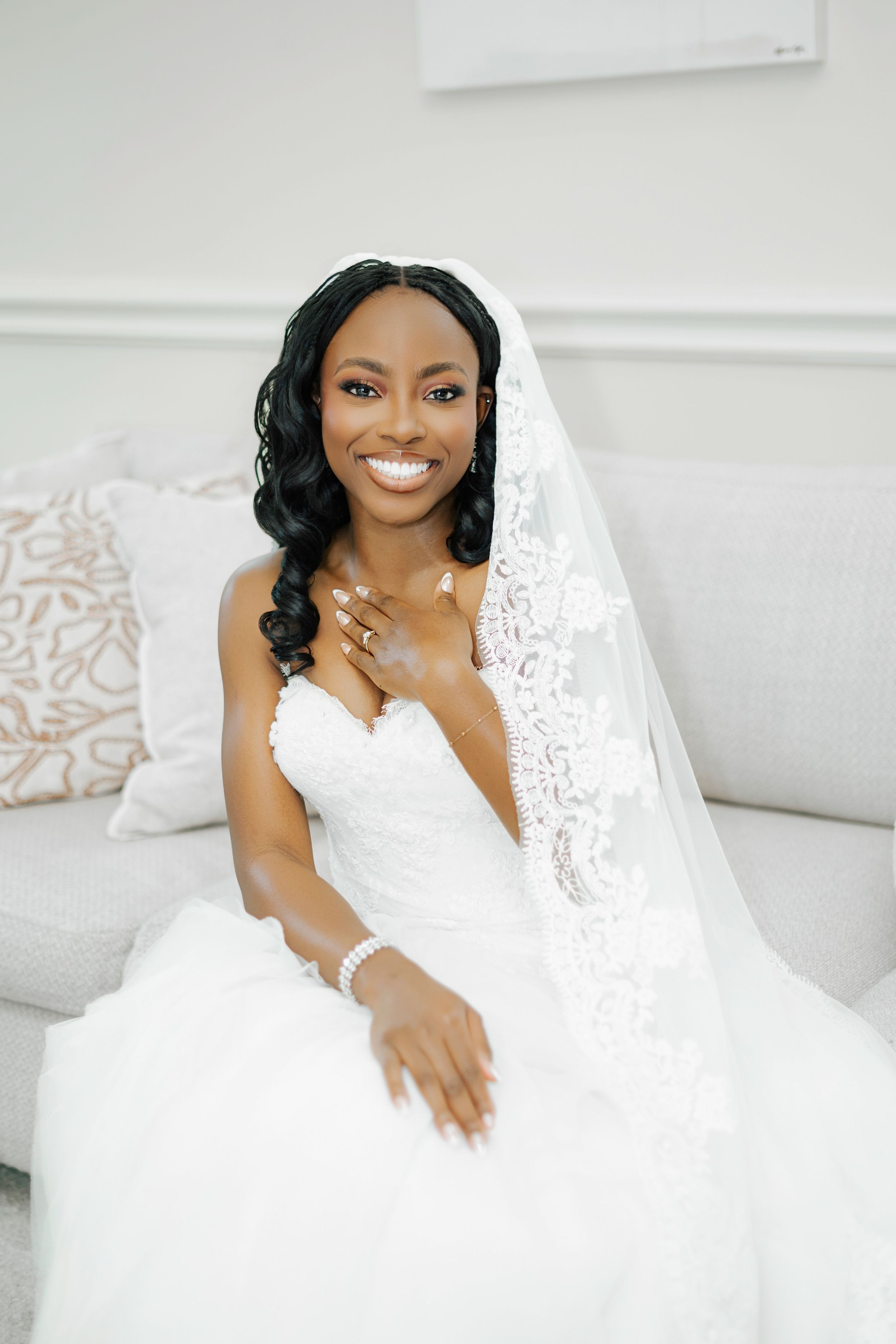 Beautiful bride in white lace wedding gown poses gracefully on white couch during bridal photoshoot.