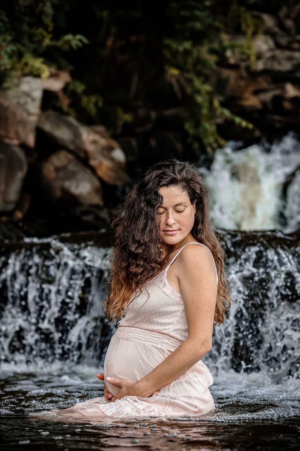 Expectant mother in white dress sitting in waterfall pool during maternity photoshoot.