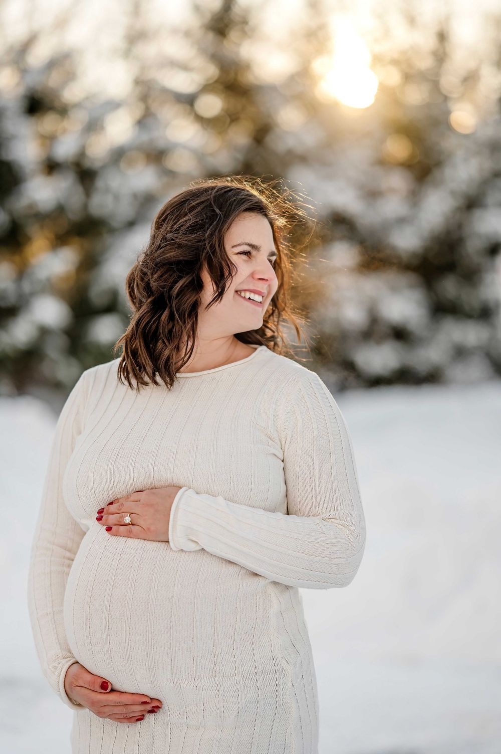 Joyful winter maternity portrait with woman in white sweater against snowy background.