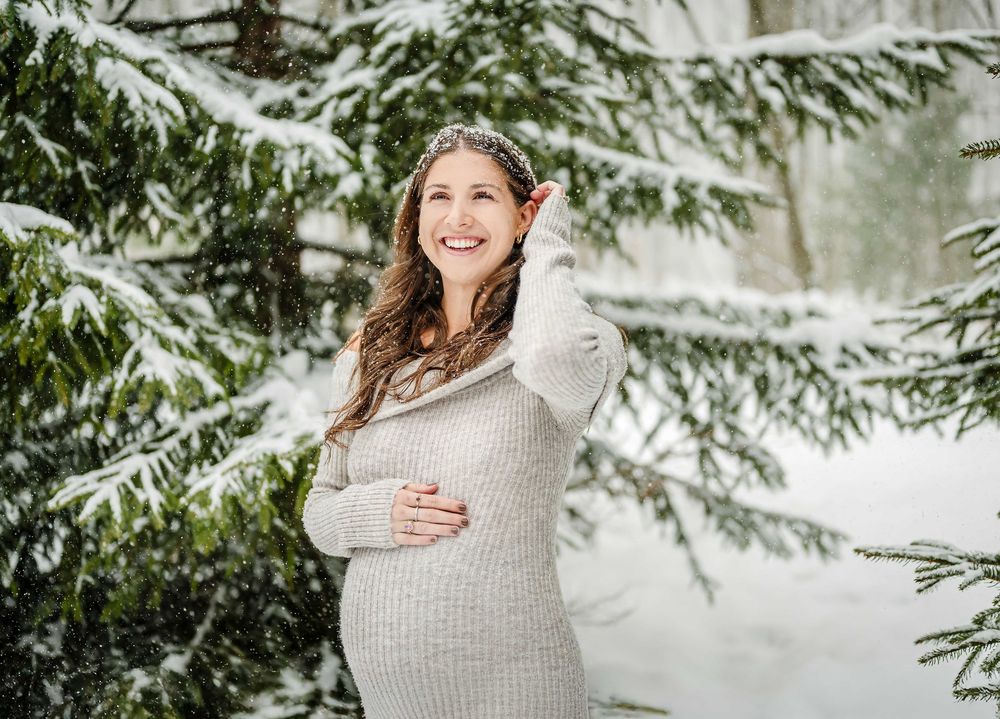 Maternity portrait of expecting mother in cream sweater surrounded by snow-covered evergreens.